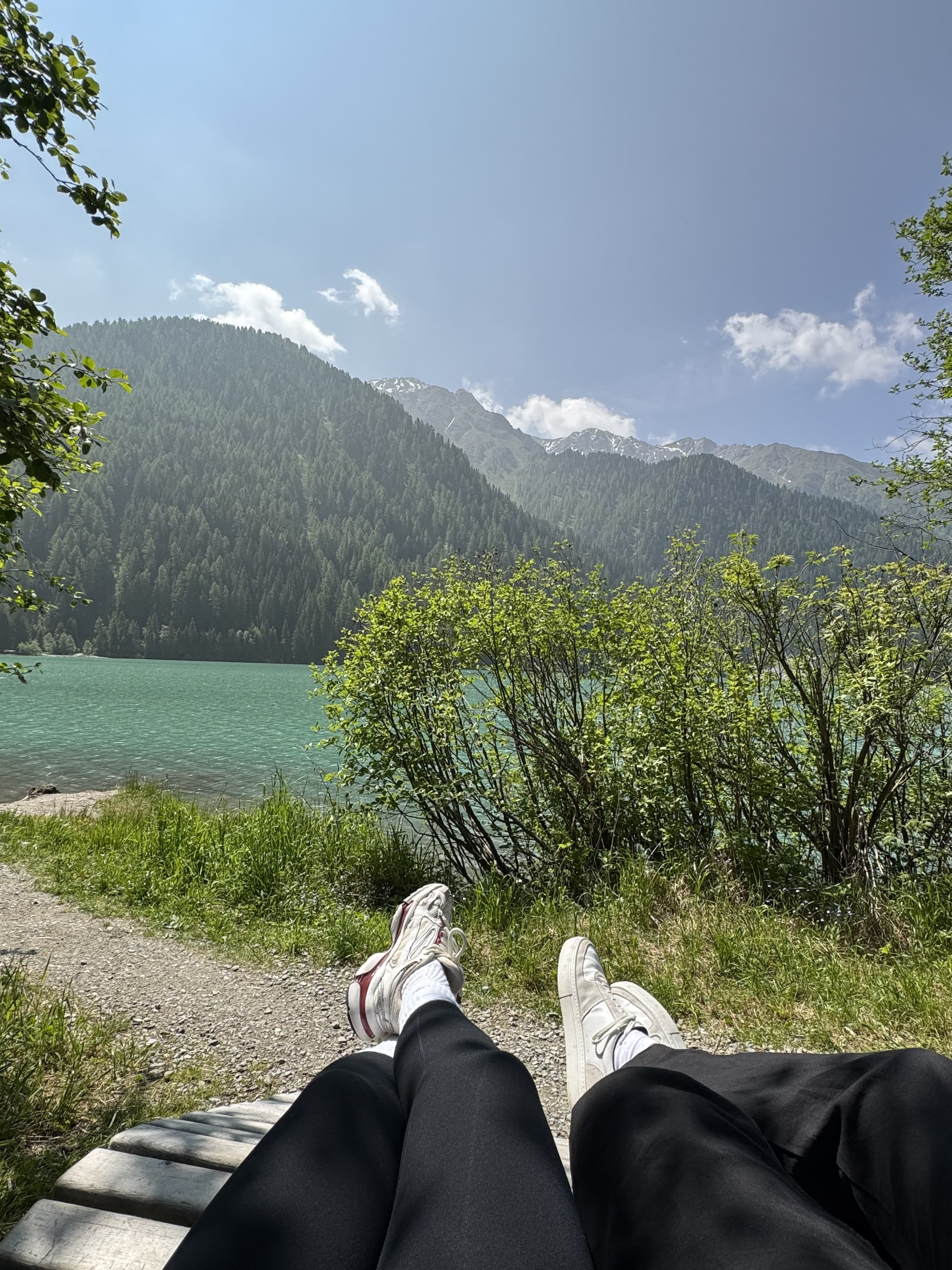Two people resting on a lakeside bench, sneakers facing turquoise water, forested mountains and blue sky with a few clouds in the background.