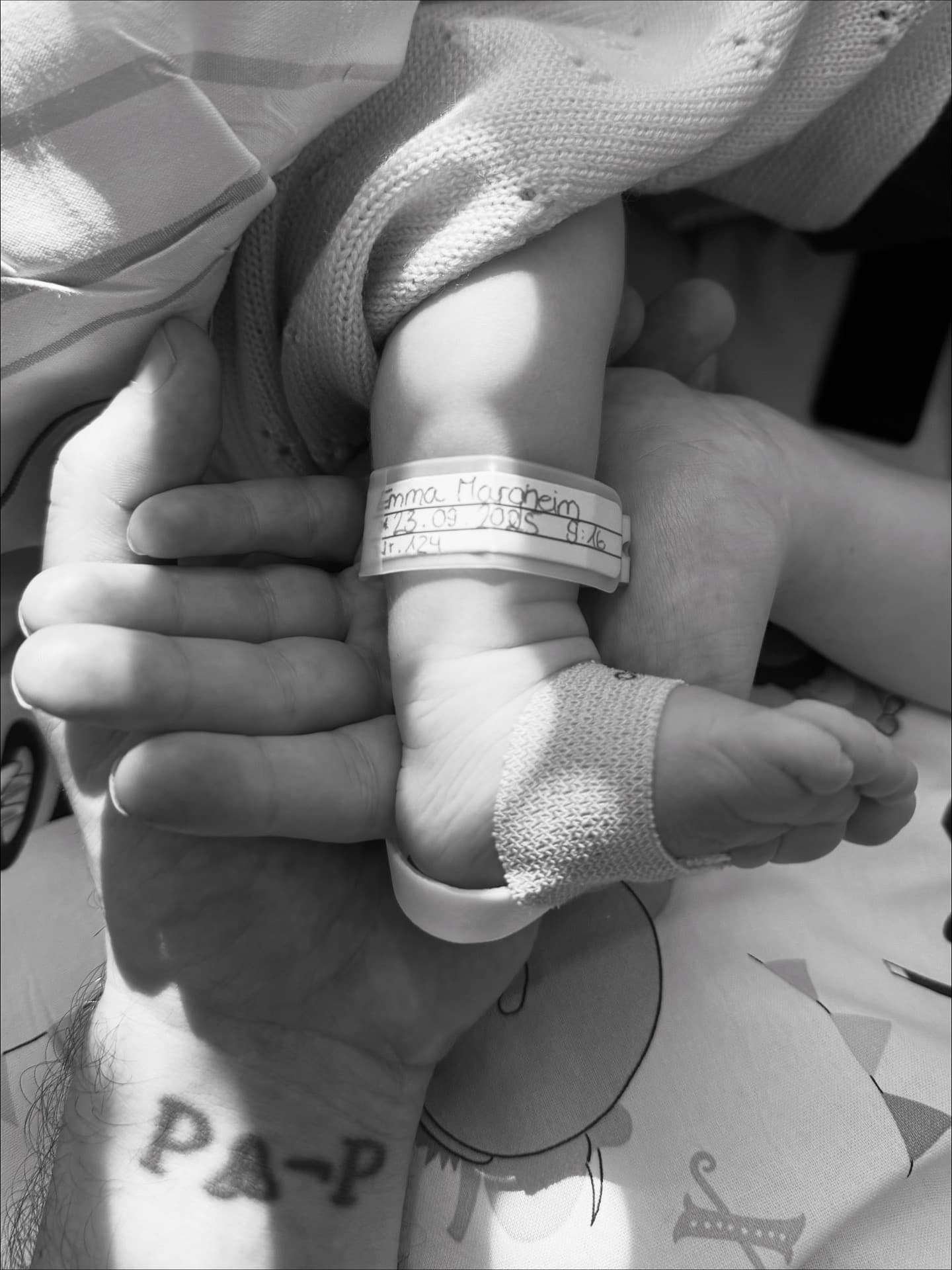 Adult hands cradling a newborn foot with hospital ID band; baby's heel has hospital tag with her name and date of birth.