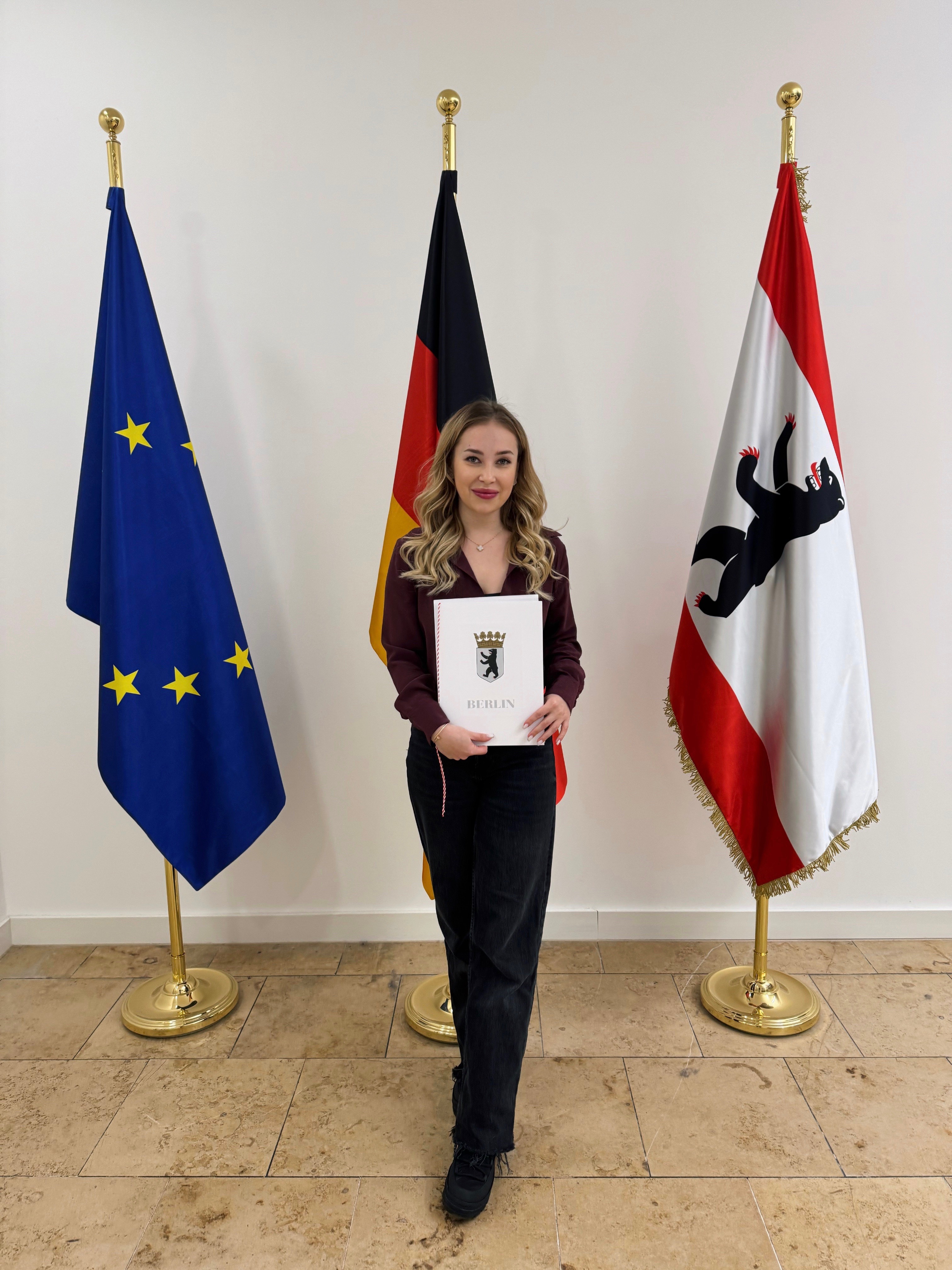 Young woman standing between European Union, German, and Berlin flags holding a Berlin certificate, smiling in an official setting.