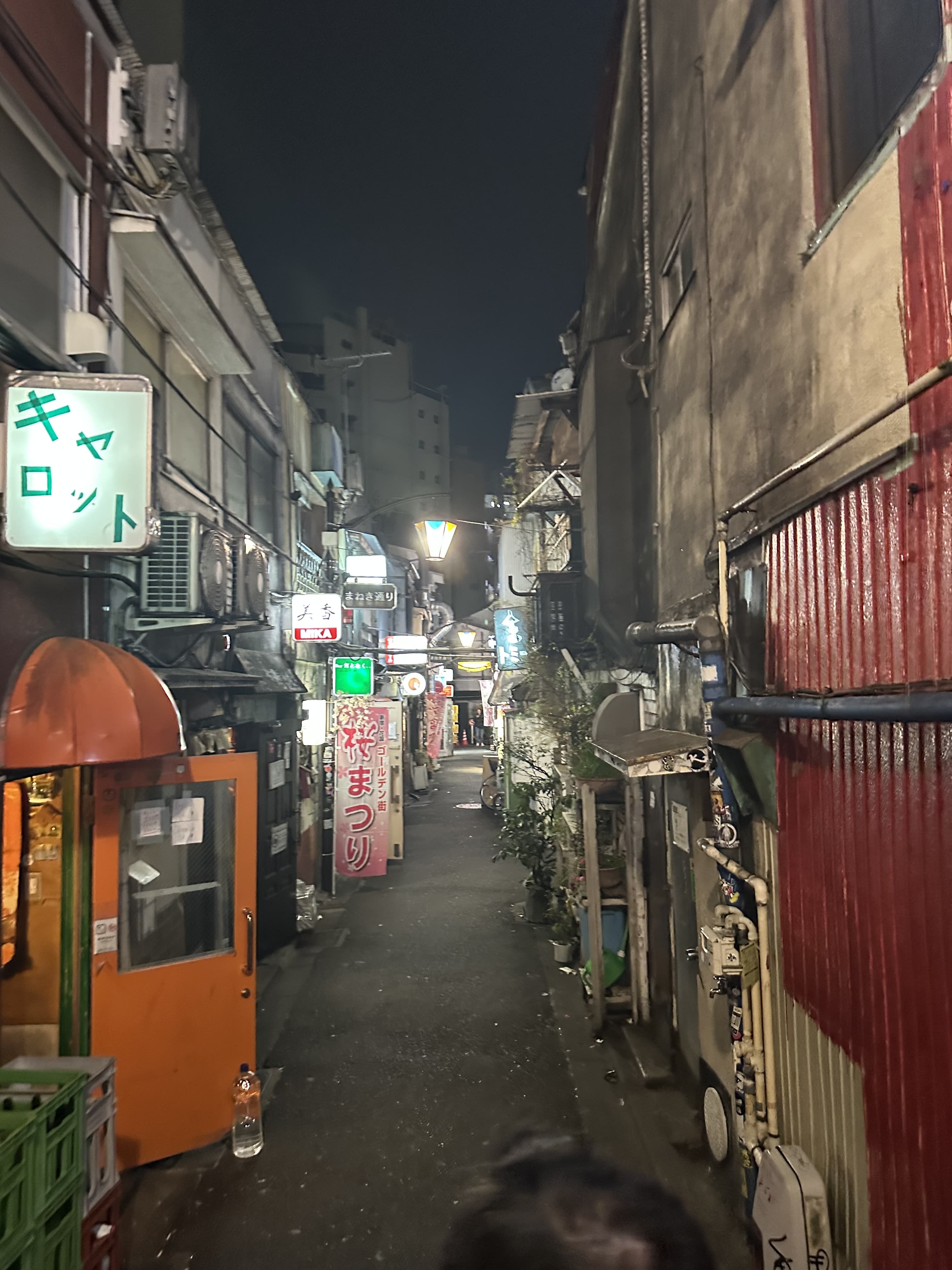 Narrow, dimly lit Japanese alley at night lined with small bars and shops, neon signs and lanterns, cluttered pipes and signage leading into distance