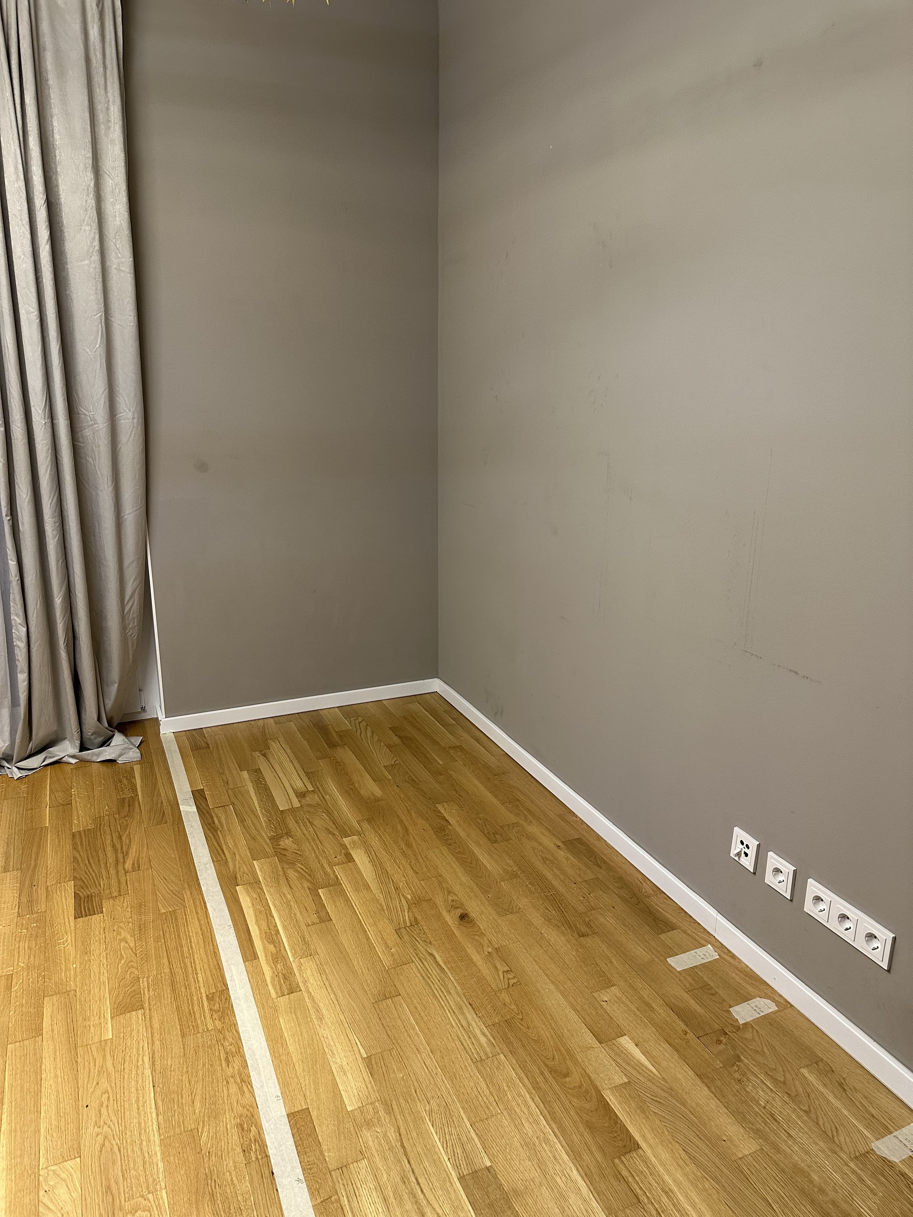 Empty corner of a room with gray walls, light wood floor, white baseboard, electrical outlets, and a gray curtain at left; painter's tape marks on floor.