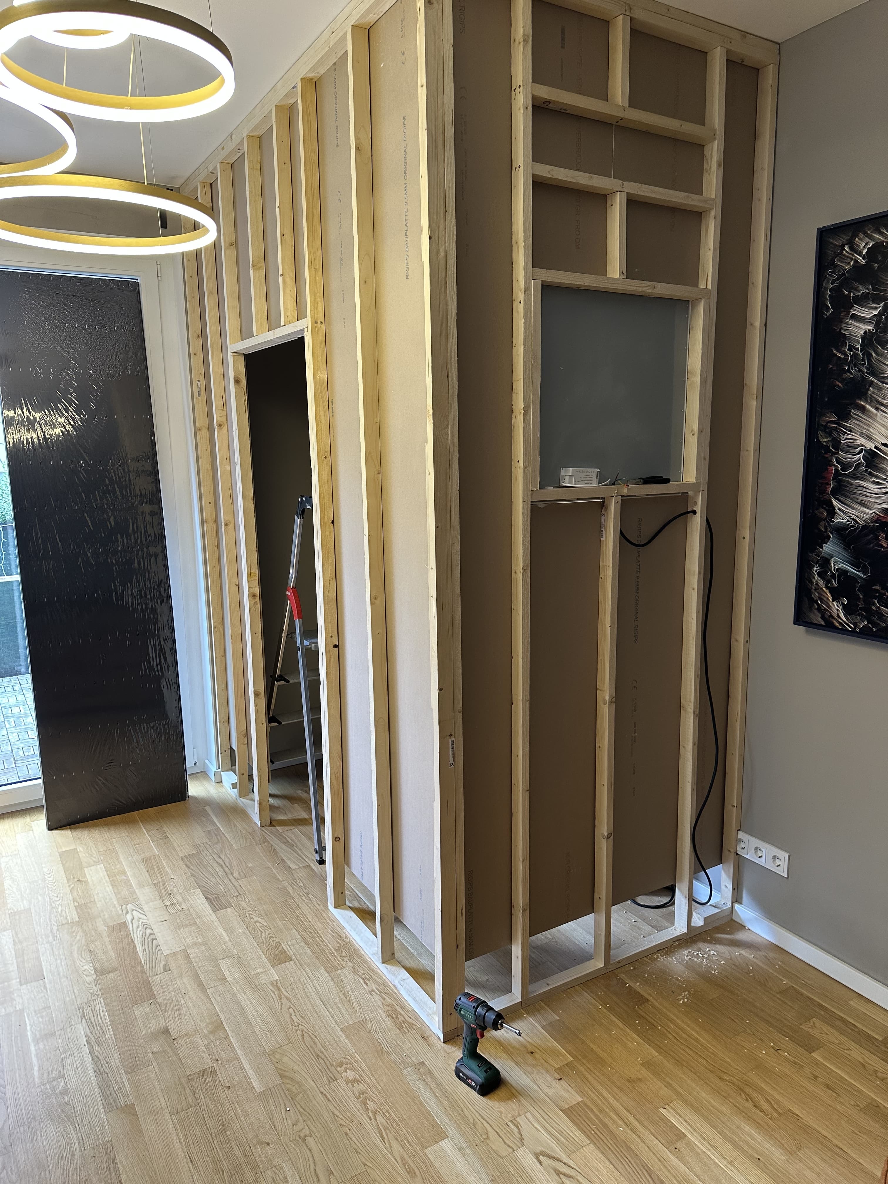 Interior room with new wooden stud framing for a partition wall, exposed wiring, a cordless drill on the floor, ladder and modern ring lights above.