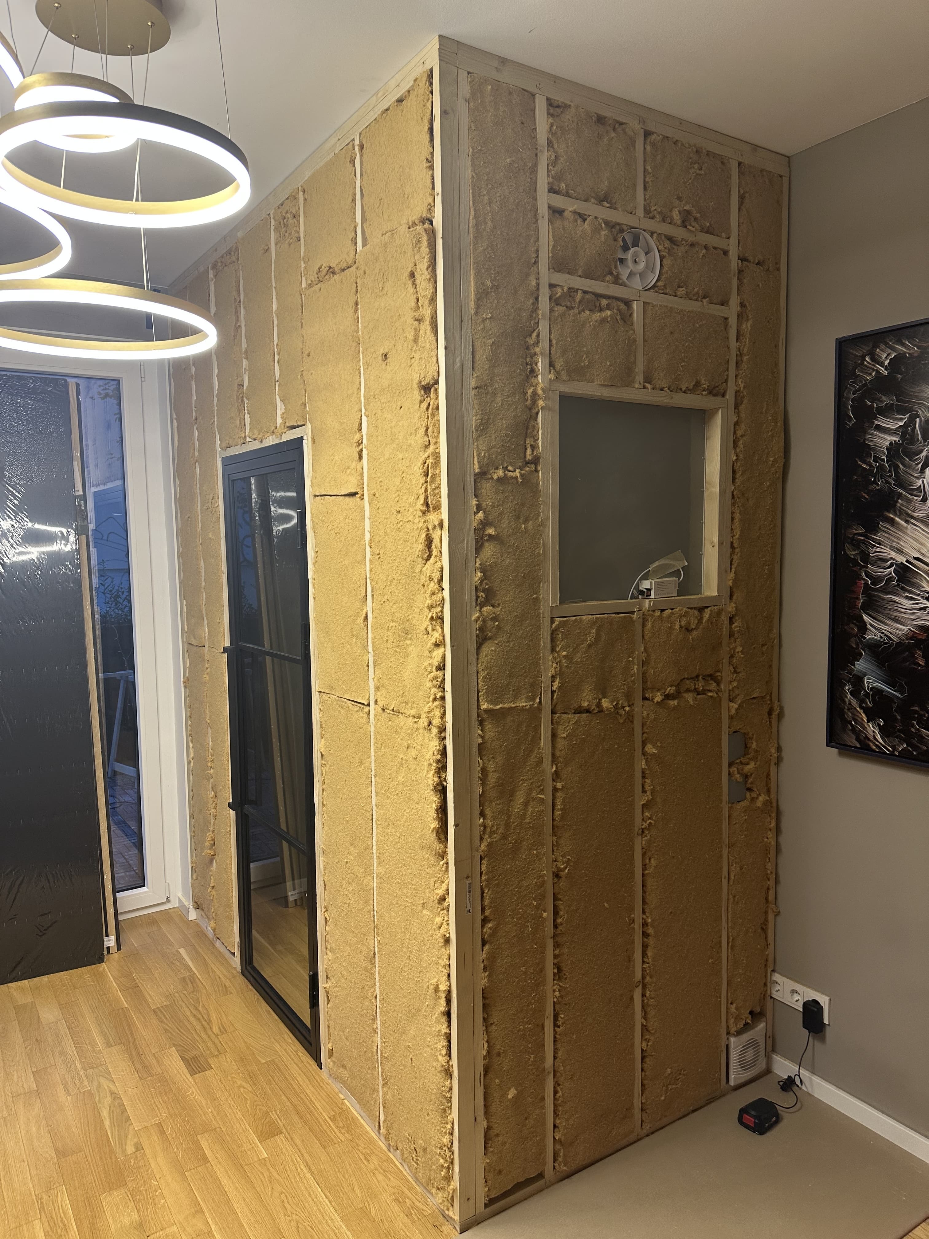 Partially built interior partition framed with insulation batts, a glass door, small window opening and ventilation fan next to hardwood flooring.