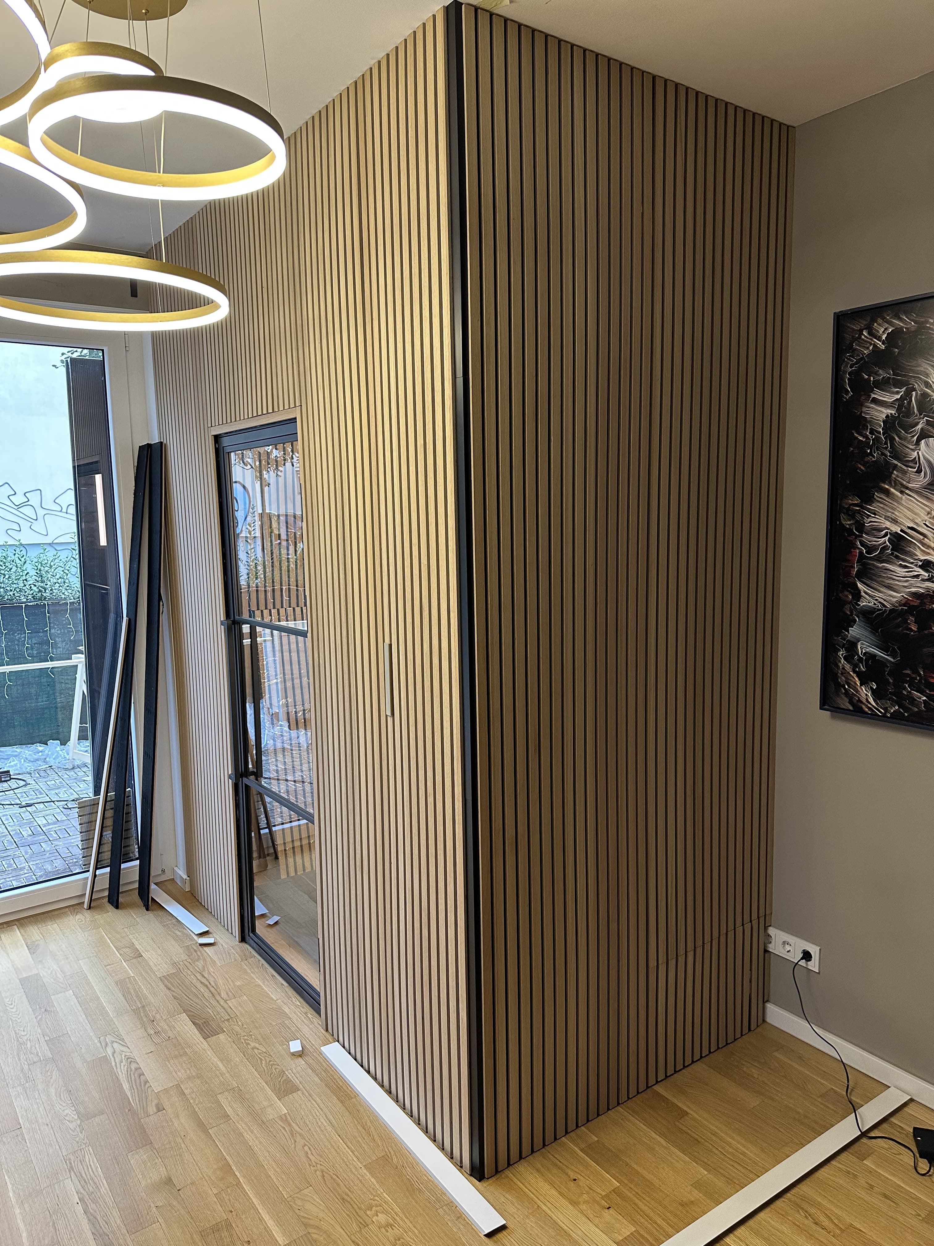 Modern room with a vertical slat wooden partition enclosing a glass-door nook, circular pendant lights overhead and hardwood floors with trim pieces on the floor.